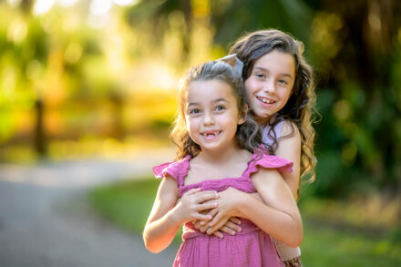 2 young sister smiling facing camera with arms wrapped around each other in a natural park setting
