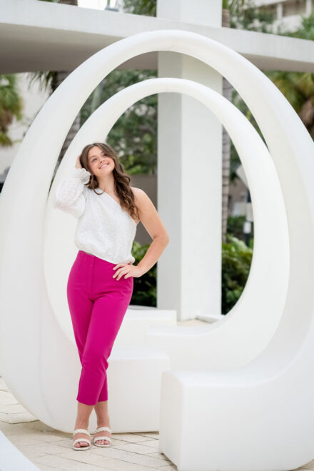 high school senior in white top and pink pants standing and smiling in front of sarasota art sculpture