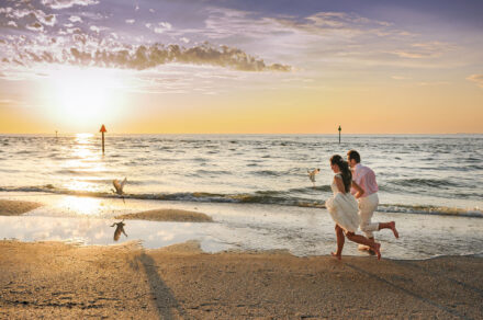 bride and groom running on beach with birds and sunset